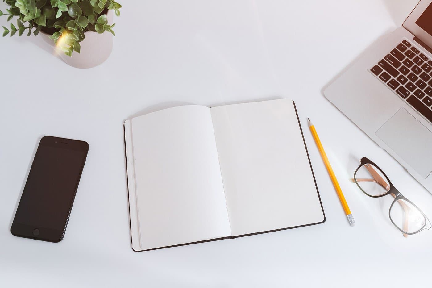 An open notebook, a pencil, glasses and a laptop on a white desk