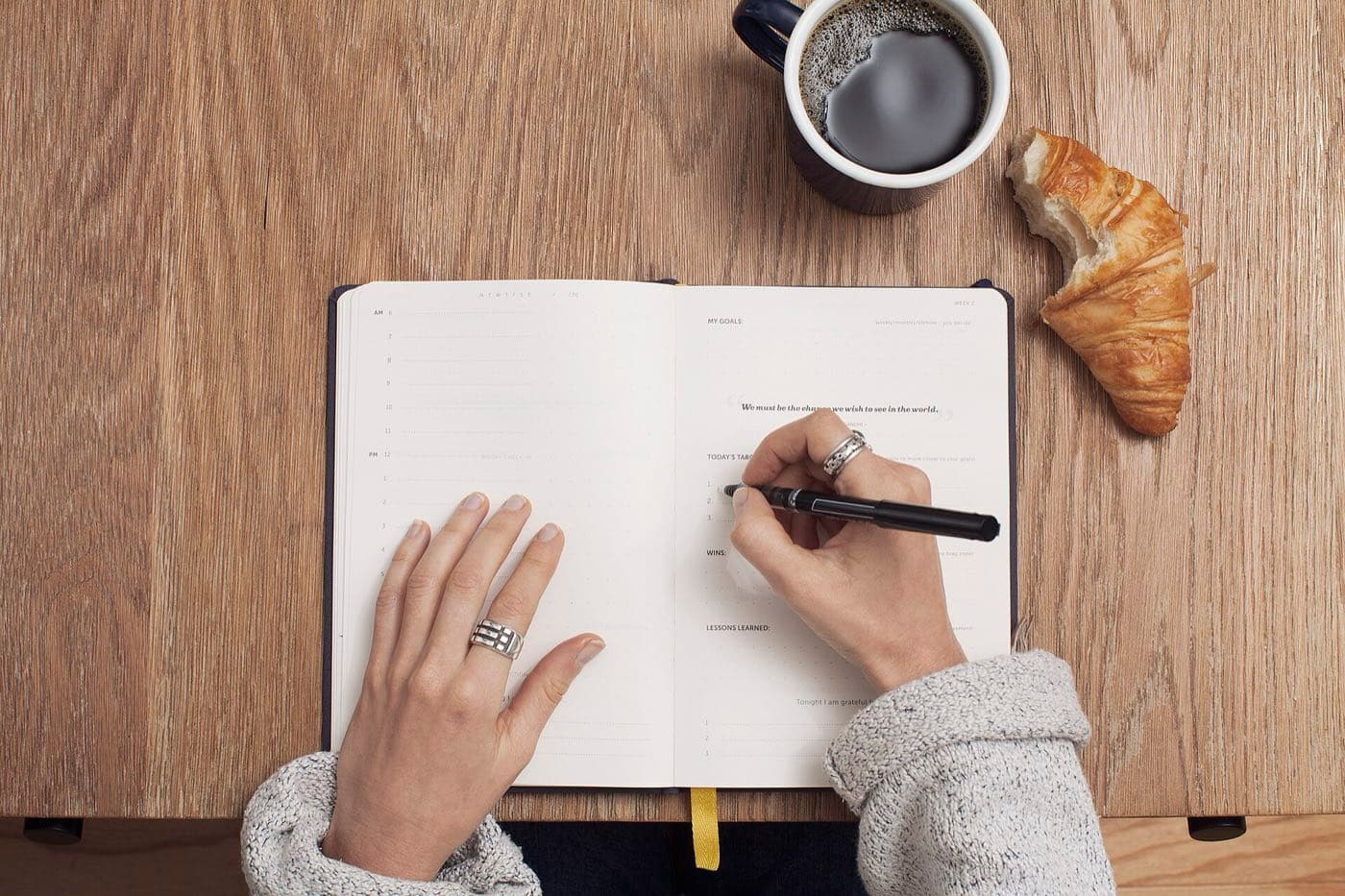 Hands writing in an open notebook beside a cup of coffee and a croissant